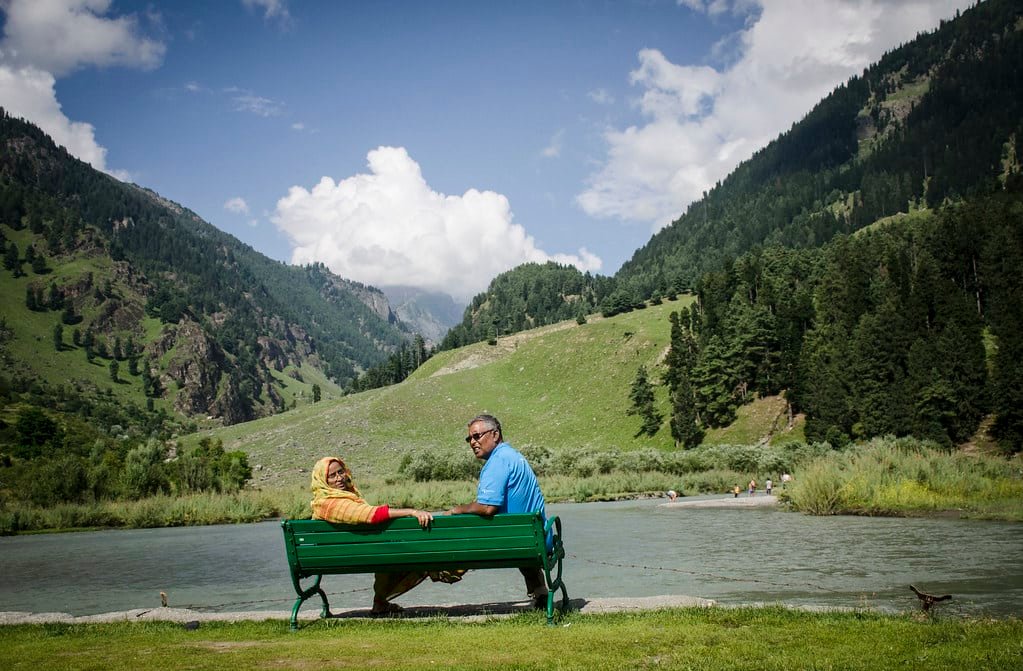 Elderly couple sitting on a bench by a river in a lush green mountain valley