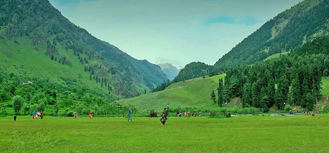 Green valley surrounded by forested mountains with people enjoying the open field.