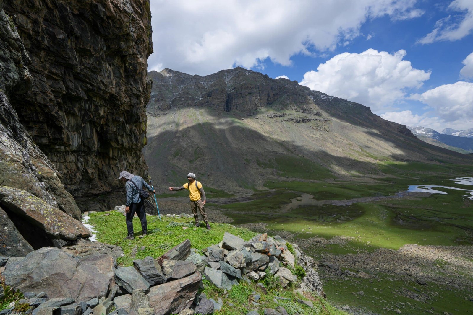 Two trekkers hike along a rocky mountain trail with lush green meadows