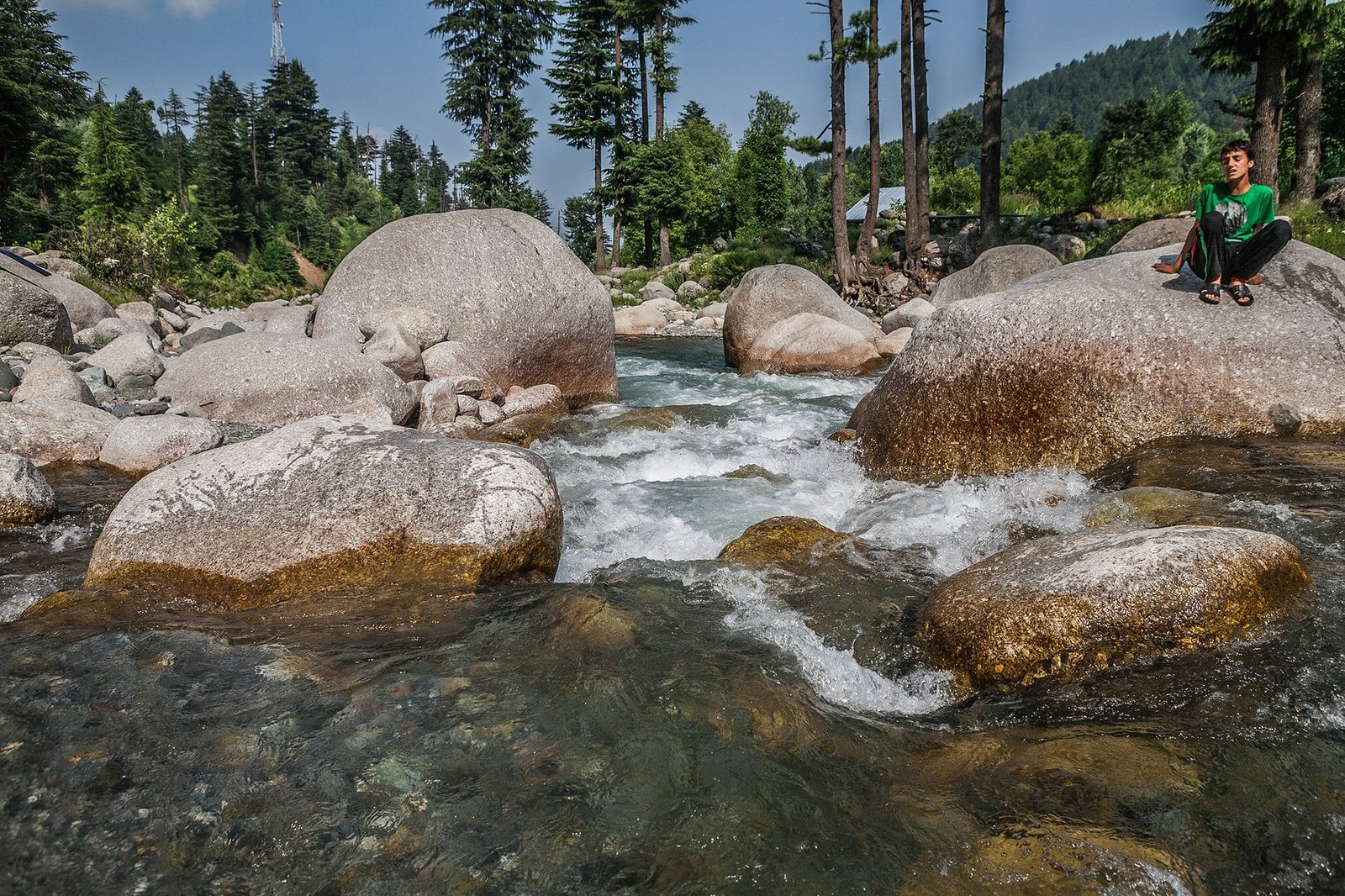 Boy sitting on a large boulder by a fast-flowing stream in a forested mountain area