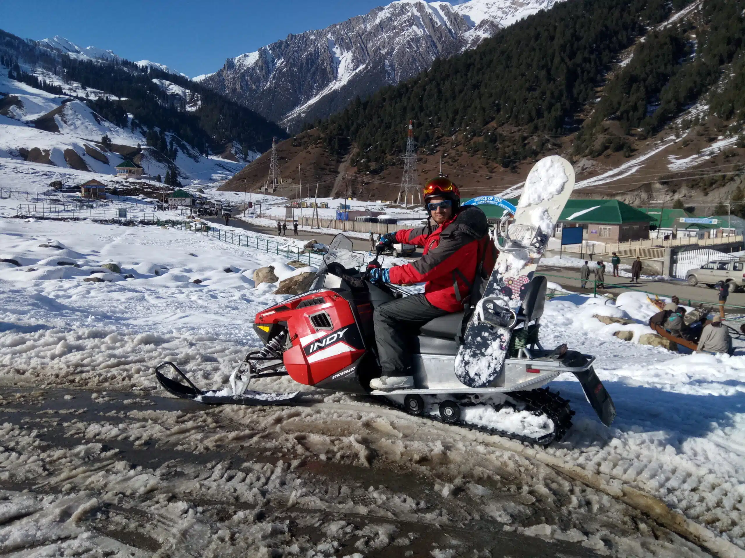 Snowmobile navigating through the snowy terrain of Gulmarg, with pine trees and Himalayan mountains in the background, showcasing winter adventure.