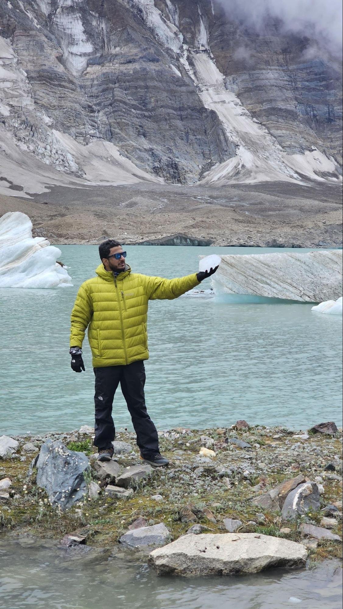 Trekker holding ice beside the lake during the Kalapari Lake Trek with glaciers and rocky mountains in the background