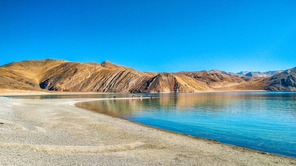 Clear blue waters of Pangong Lake with surrounding brown mountains under a bright sky - one of the most stunning places to visit in Leh.