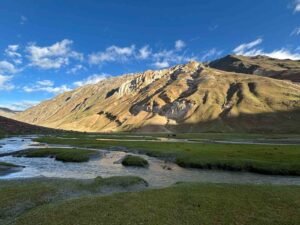 A serene river winds through lush green meadows beneath sunlit mountains, showcasing the vibrant beauty of Ladakh in May.